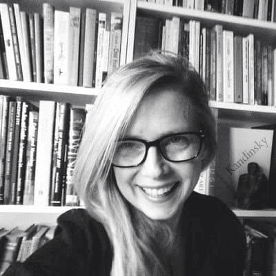 A black and white photo of a smiling woman with glasses in front of a bookshelf.
