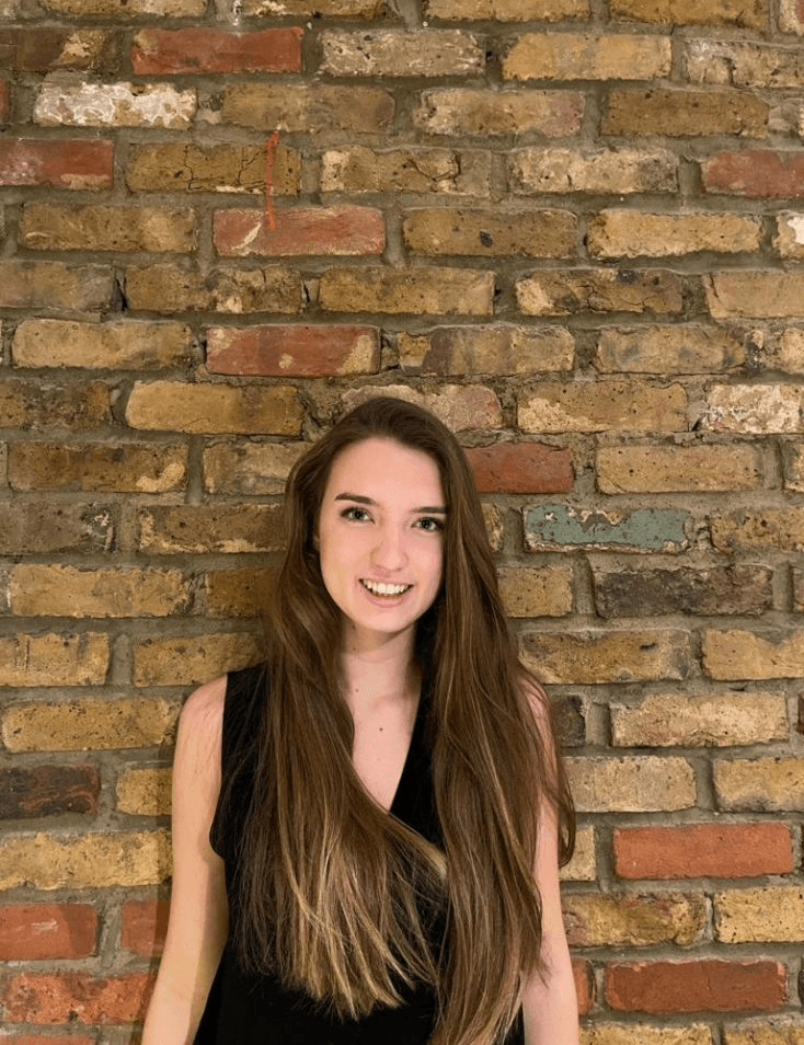 A smiling young woman with long brown hair standing in front of a brick wall.