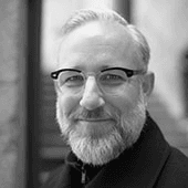 A black and white portrait of a smiling man with a beard wearing glasses.