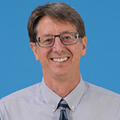 A smiling middle-aged man with glasses, wearing a light blue shirt and a dark blue tie, standing against a blue background.