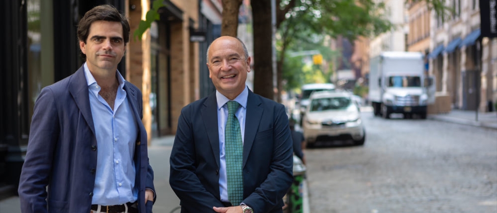 Two men in business attire walking on a city street with one looking at the camera and the other looking away.