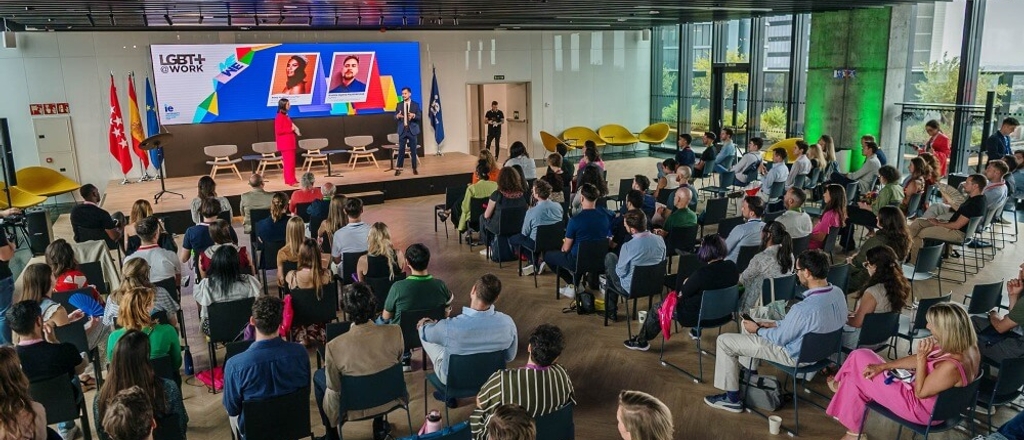 A conference setting with attendees seated observing a presentation at a brightly lit venue with prominent displays showing LGBT+ flags and supportive messages.