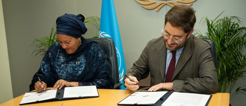 Two individuals, one woman and one man, are signing documents at a desk with a UN logo visible in the background.