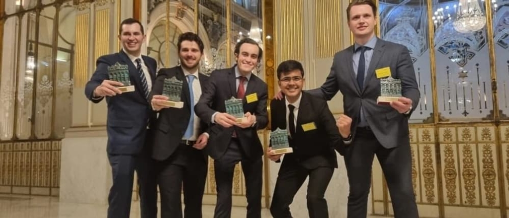 Five men in formal attire holding books and smiling in a grand hallway.