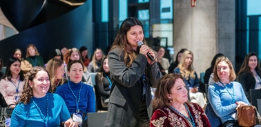 A woman stands up to speak at a conference while others listen attentively in the audience.