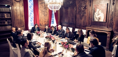 A formal meeting taking place in an elegant, wood-paneled room with a large chandelier and a modern painting.