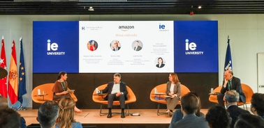 A panel discussion taking place at IE University with four speakers and a large screen displaying the Amazon logo.
