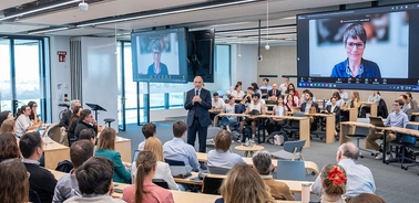 A speaker addresses an audience in a conference room while projected on a screen is another participant.