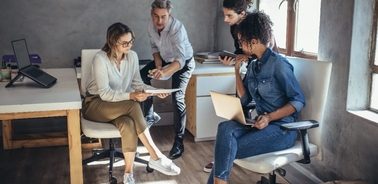 Four colleagues collaborate in a modern office, discussing ideas while using laptops and tablets.