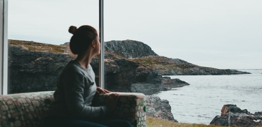 A person sitting by a window, gazing at a rocky seascape on a cloudy day.