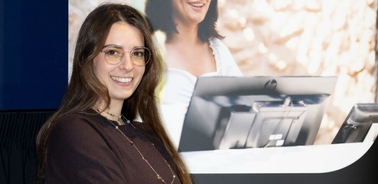 A woman in glasses standing at a presentation podium with a projected image of another woman in the background.