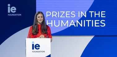 A woman in a red dress standing at a podium with a sign 'IE Foundation' and text 'PRIZES IN THE HUMANITIES' in the background.