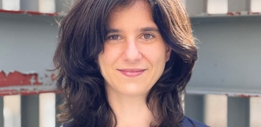 A portrait of a smiling woman with shoulder-length dark hair standing in front of a metal gate.