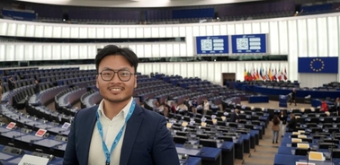 A man smiling in the European Parliament chamber with flags and seats in the background.