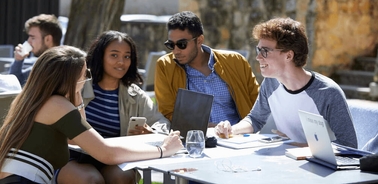 A group of young adults engaging in a discussion at an outdoor table, with laptops and smartphones visible.