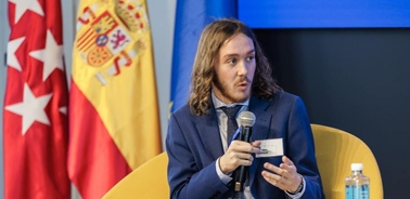 A man with long hair speaks into a microphone at a panel discussion, with Spanish and European Union flags in the background.