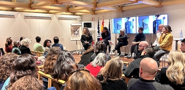 A diverse audience engaged in a discussion during an event with speakers on stage.