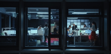View of a busy office through the windows at night, with multiple people working at their desks.