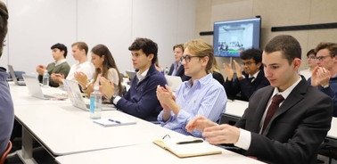A group of young adults in a classroom applauding, some dressed in formal attire.