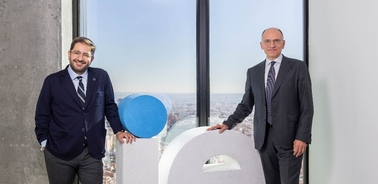 Two men in business suits standing beside a large letter 'Q' in a modern office setting overlooking a cityscape.