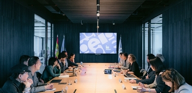 A group of people engaged in a meeting around a long table with a large screen displaying graphics.