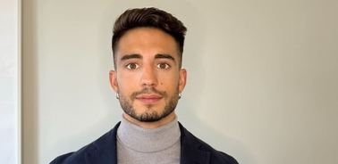 A young man with styled hair, wearing a turtleneck and a blazer, standing indoors.