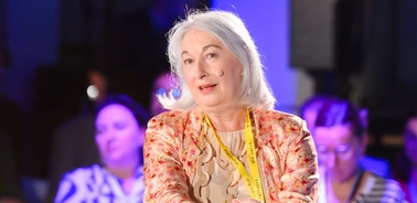 An older woman with white hair sits attentively, wearing a bright floral blazer over a white blouse.