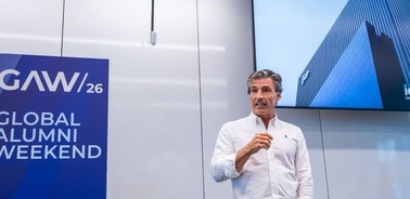 A speaker presenting at the Global Alumni Weekend event with a large screen displaying a modern building.