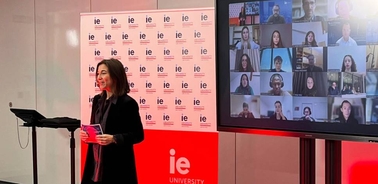 A woman stands speaking at a podium in front of a branded IE University backdrop, with a screen showing multiple virtual attendees behind her.