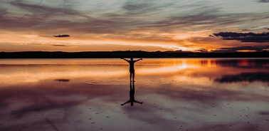 A person with arms spread wide stands in shallow water under a vivid sunset sky.