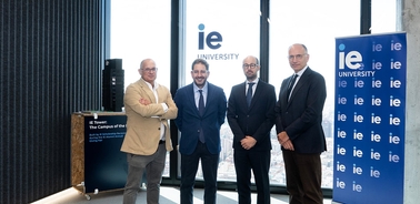 Four professionals pose together in an urban setting with the IE University logo in the background.