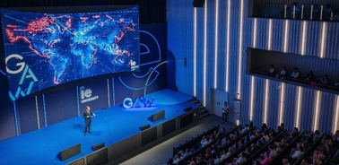 A speaker presenting at a conference with a large digital screen displaying a global map in the background.