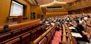 A scene in a lecture hall with people listening to a presenter at the front.