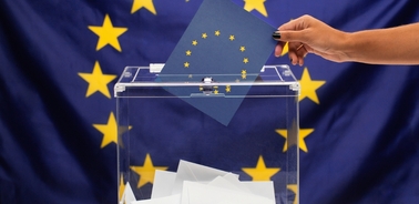 A hand placing a ballot into a transparent ballot box in front of an EU flag.