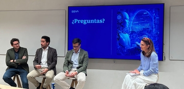 A panel discussion with four people on stage, including a woman taking notes and a large screen displaying a question.