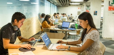 A young man and woman are working on laptops in a busy office environment.