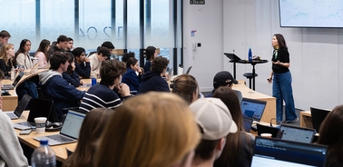 A university classroom lecture with a female professor presenting to a group of students seated at desks.