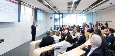 A speaker presenting to a group of attentive professionals in a modern conference room.