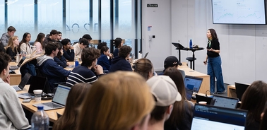 A professor is teaching a class filled with students seated at desks with laptops.