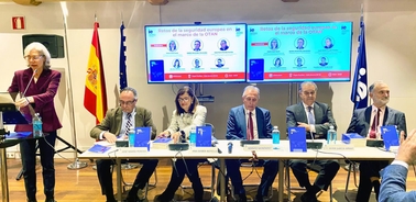 A professional conference with seven individuals seated at a panel in front of flags and presentation screens.