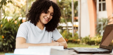 A young woman with curly hair smiling while studying outdoors with a laptop and books on the table.