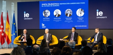 Several men are sitting on stage at a panel discussion, with flags and presentation screens in the background.