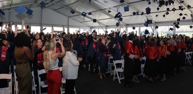Graduates joyfully tossing their caps in the air during a graduation ceremony under a large tent.