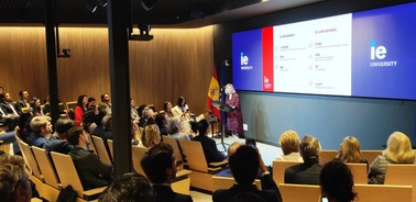 A woman presenting at a business conference in front of an audience, with a digital presentation screen displaying graphics behind her.