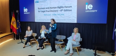 A woman is speaking at a podium during a business and human rights forum at IE University, while three participants sit nearby listening.