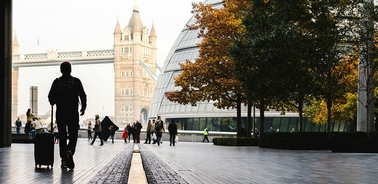 A silhouette of a person with a suitcase walking in an urban area with trees and modern buildings.