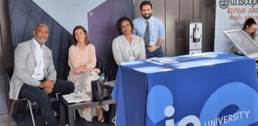 Four individuals, two men and two women, are seated and standing around a promotional booth with a 'IE University' banner.