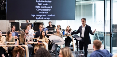 A man is presenting in front of a group of seated young adults in a modern seminar room.