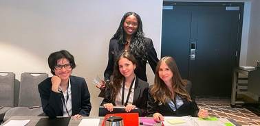 A group of four women in professional attire are seated at a table in a conference setting.