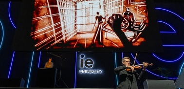 A musician plays the violin on stage while an artistic performance is displayed on a large screen behind him.